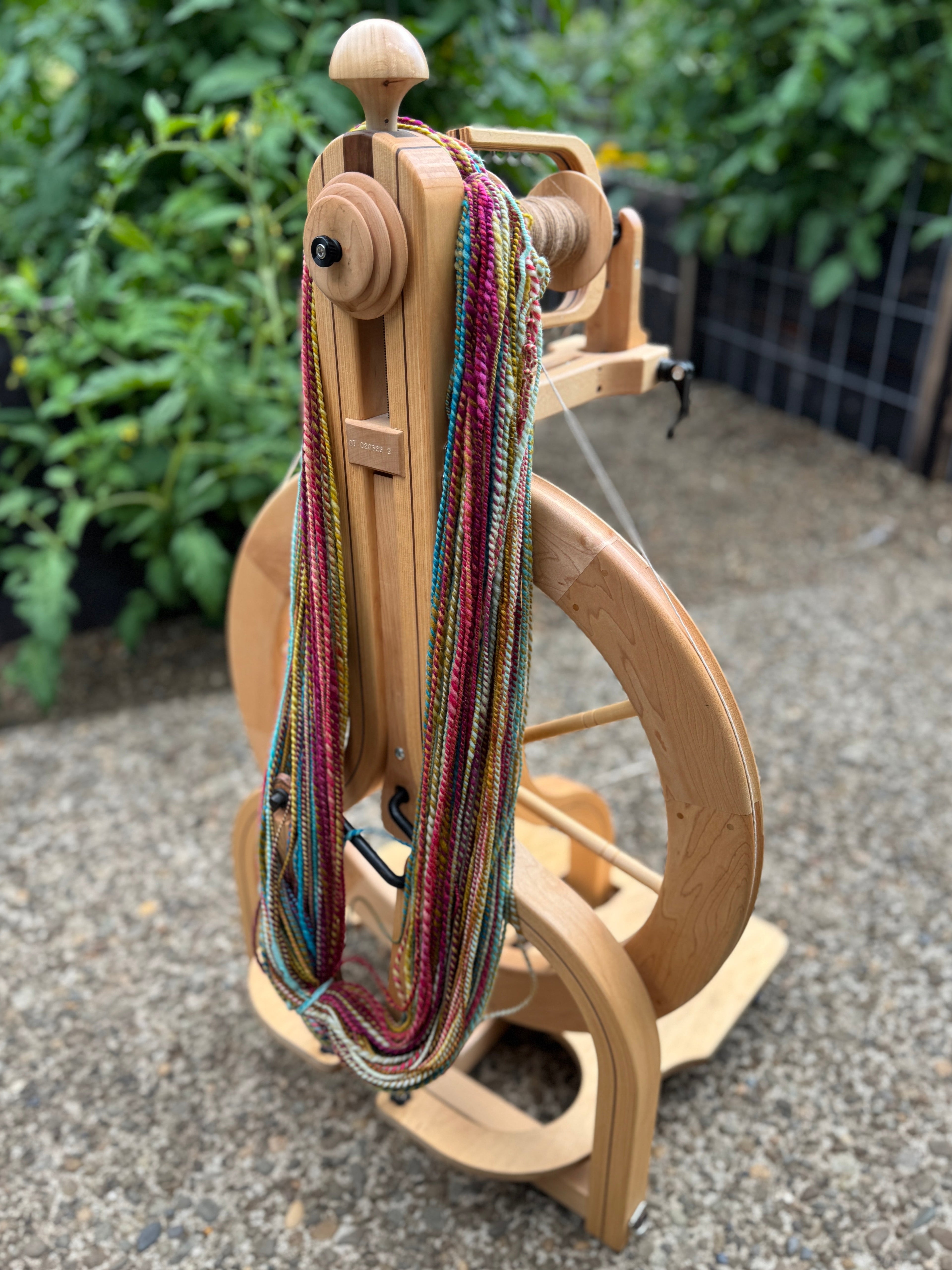 Wooden spinning wheel with colorful yarn on a gravel surface with greenery in the background